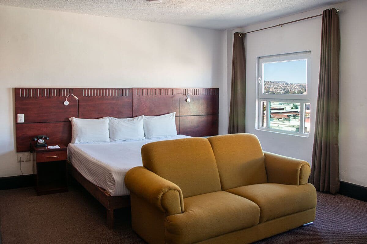 Habitación de hotel con cama, sofá, mesita de noche, lámpara y ventana con vistas.