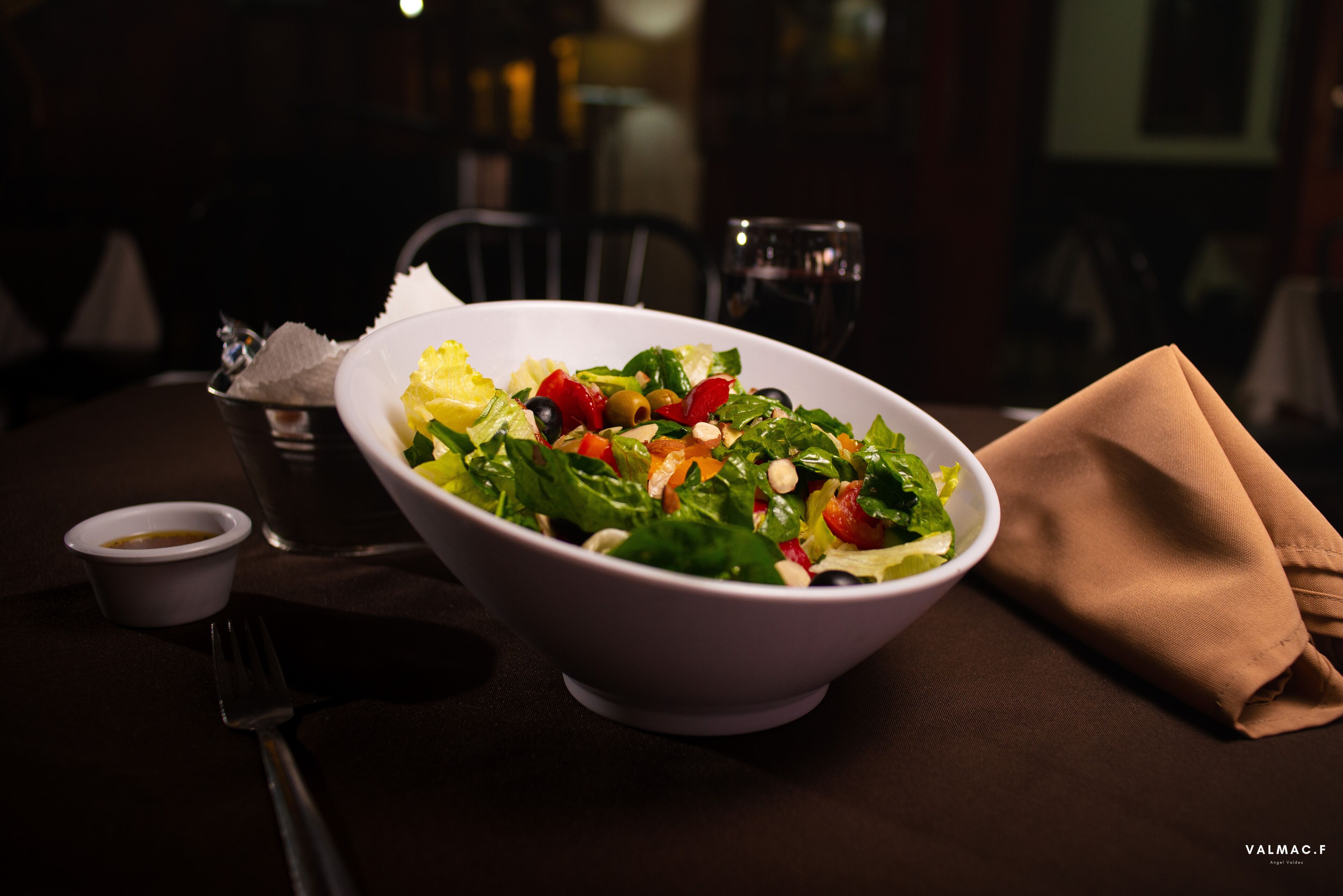 A white bowl of salad with various vegetables on a table, accompanied by a small cup of dressing and a folded napkin.