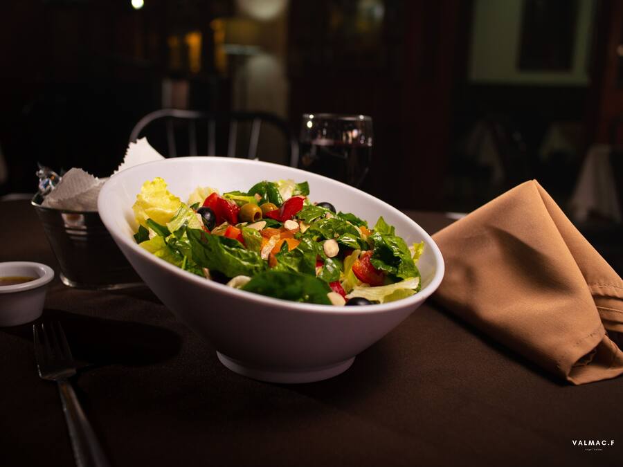 A white bowl of salad with various vegetables on a table, accompanied by a small cup of dressing and a folded napkin.