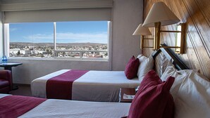 A hotel room with two beds, a view of a cityscape through a large window, and a wooden wall.