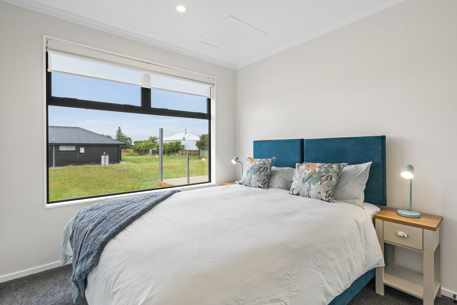 A bedroom with a large window, a bed with a blue headboard, two bedside lamps, and a view of a building and greenery outside.