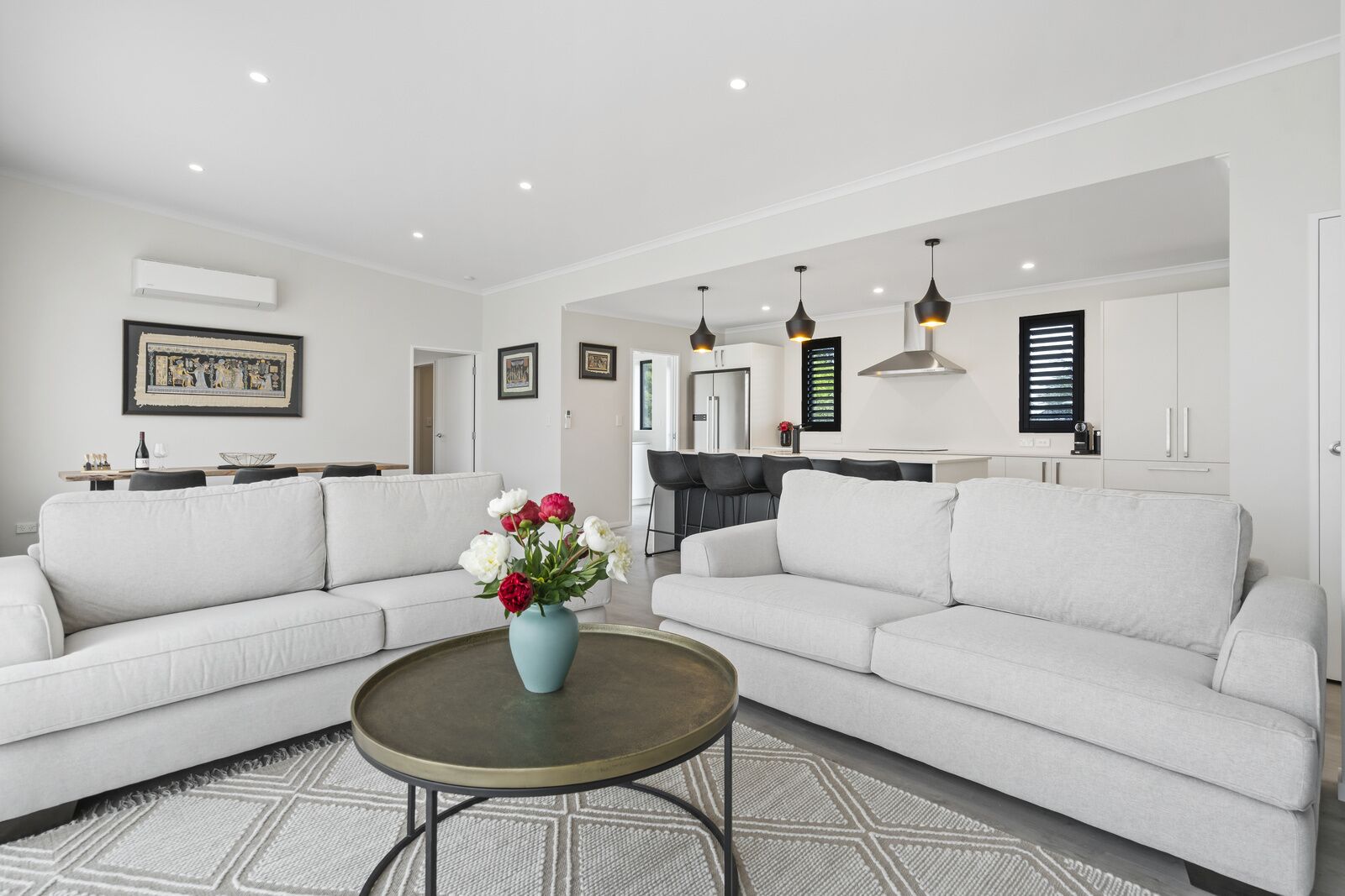 Una sala de estar moderna con dos sofás blancos, una mesita de centro y un jarrón con flores. Se observa una cocina al fondo con una mesa de comedor y sillas.