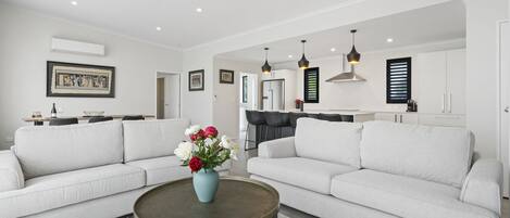 Una sala de estar moderna con dos sofás blancos, una mesita de centro y un jarrón con flores. Se observa una cocina al fondo con una mesa de comedor y sillas.
