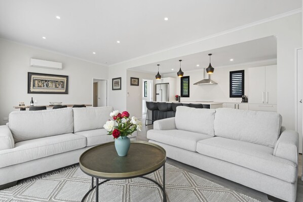 Una sala de estar moderna con dos sofás blancos, una mesita de centro y un jarrón con flores. Se observa una cocina al fondo con una mesa de comedor y sillas.