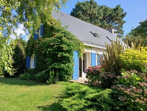 A house with a sloped roof, blue shutters, and a garden with various plants and flowers.