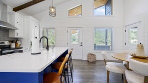 A modern kitchen with white cabinetry, a large island with bar stools, a wooden dining table with chairs, and a view of the outdoors through windows.