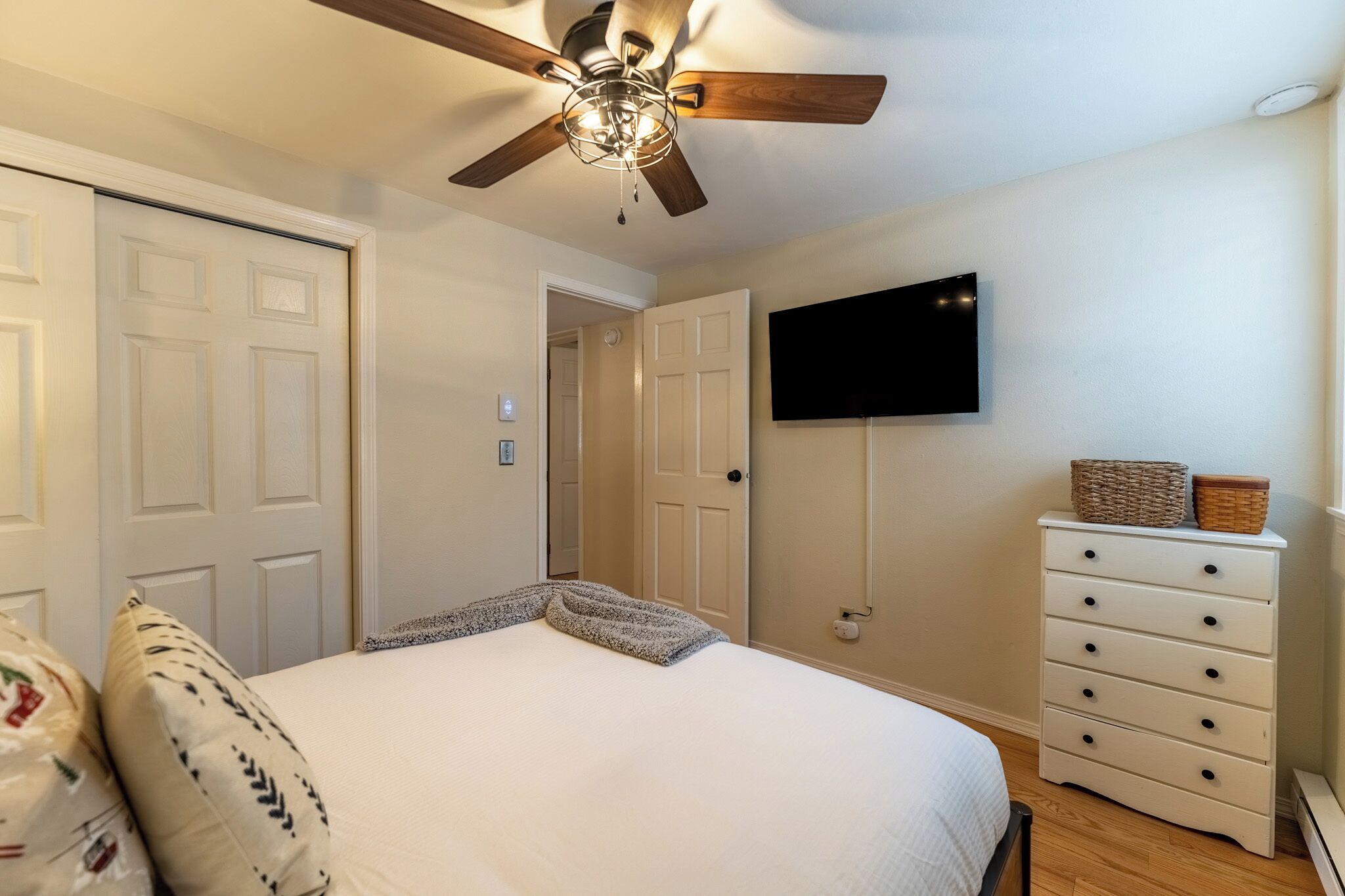 A bedroom with a bed, a ceiling fan, a TV mounted on the wall, and a dresser with baskets.