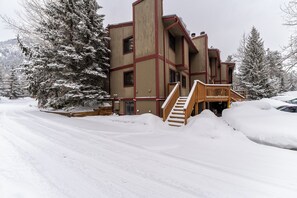 A multi-story building with a wooden staircase leading to the entrance, surrounded by snow-covered trees and parked cars.