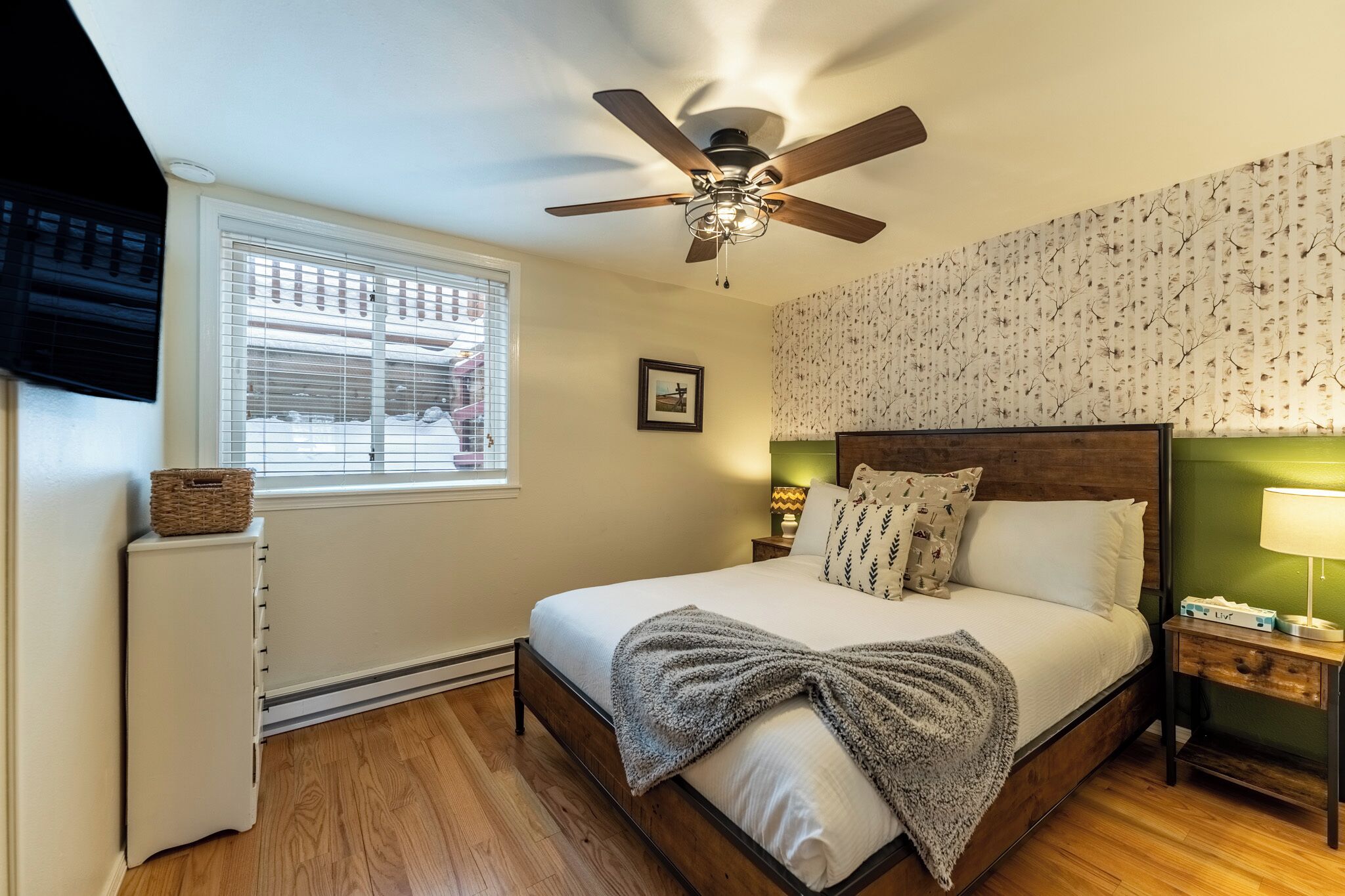 Un dormitorio con cama, ventilador de techo, mesita de noche, lámpara, ventana con persianas y radiador.