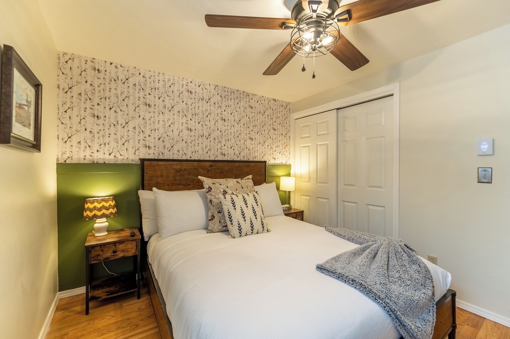 Un lit bien fait, avec une tête de lit en bois, une table de chevet avec une lampe, un ventilateur de plafond et une porte avec un interrupteur.