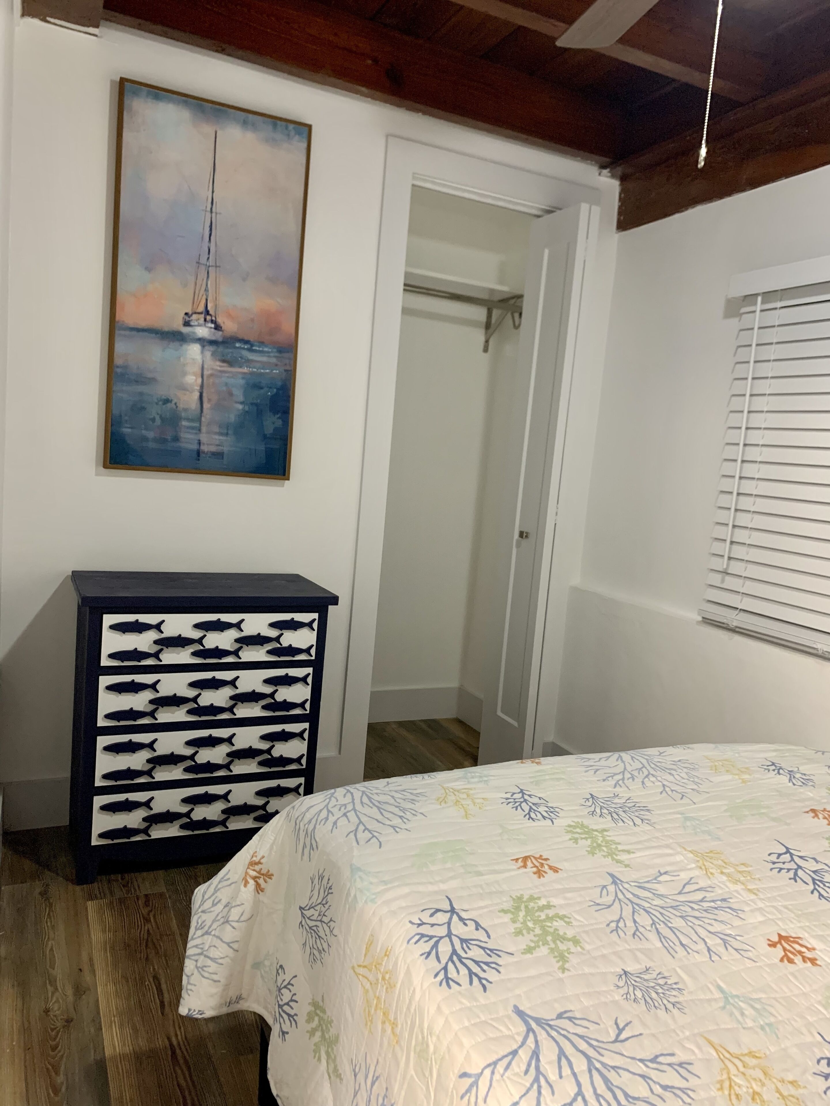 Een slaapkamer met een bed, een dressoir met vissen uitsparingen, een schilderij van een zeilboot en een kast.