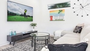A modern living room with a flat-screen TV, a round glass coffee table, a sofa, and a wall clock. - Golfer's Paradise: EV Charger, Private Pool & Putting (La Quinta)