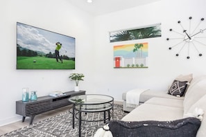 A modern living room with a flat-screen TV, a round glass coffee table, a sofa, and a wall clock. - Golfer's Paradise: EV Charger, Private Pool & Putting (La Quinta)