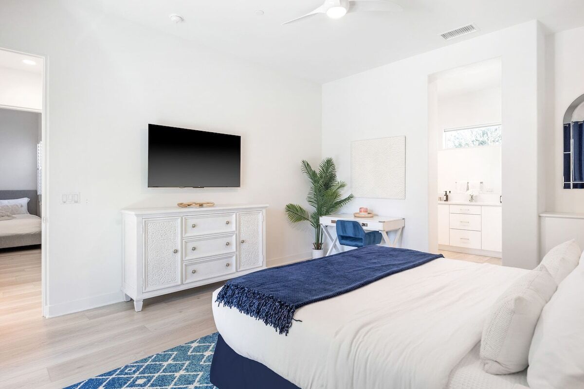 Quarto com cama, televisão presa na parede, escrivaninha com cadeira e banheiro visível através de uma porta aberta.