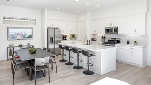 A modern kitchen with white cabinetry, a central island with bar stools, a dining area with a table and chairs, and a view of the outdoors through a window. - Golfer's Paradise: EV Charger, Private Pool & Putting (La Quinta)