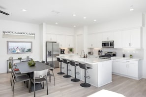 A modern kitchen with white cabinetry, a central island with bar stools, a dining area with a table and chairs, and a view of the outdoors through a window. - Golfer's Paradise: EV Charger, Private Pool & Putting (La Quinta)
