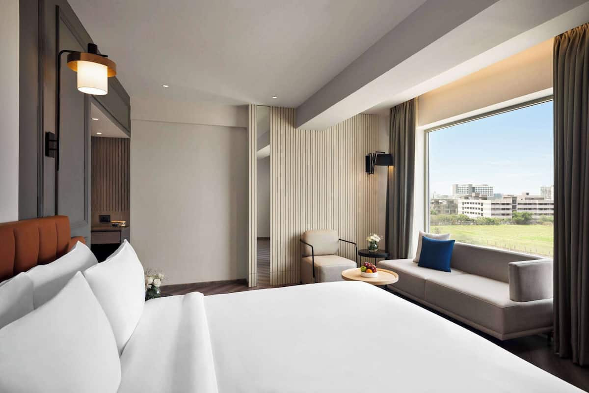Quarto de hotel moderno com uma cama grande, um sofá, uma mesinha com uma cesta de frutas, uma cadeira, uma TV e uma janela ampla com vista para a cidade.