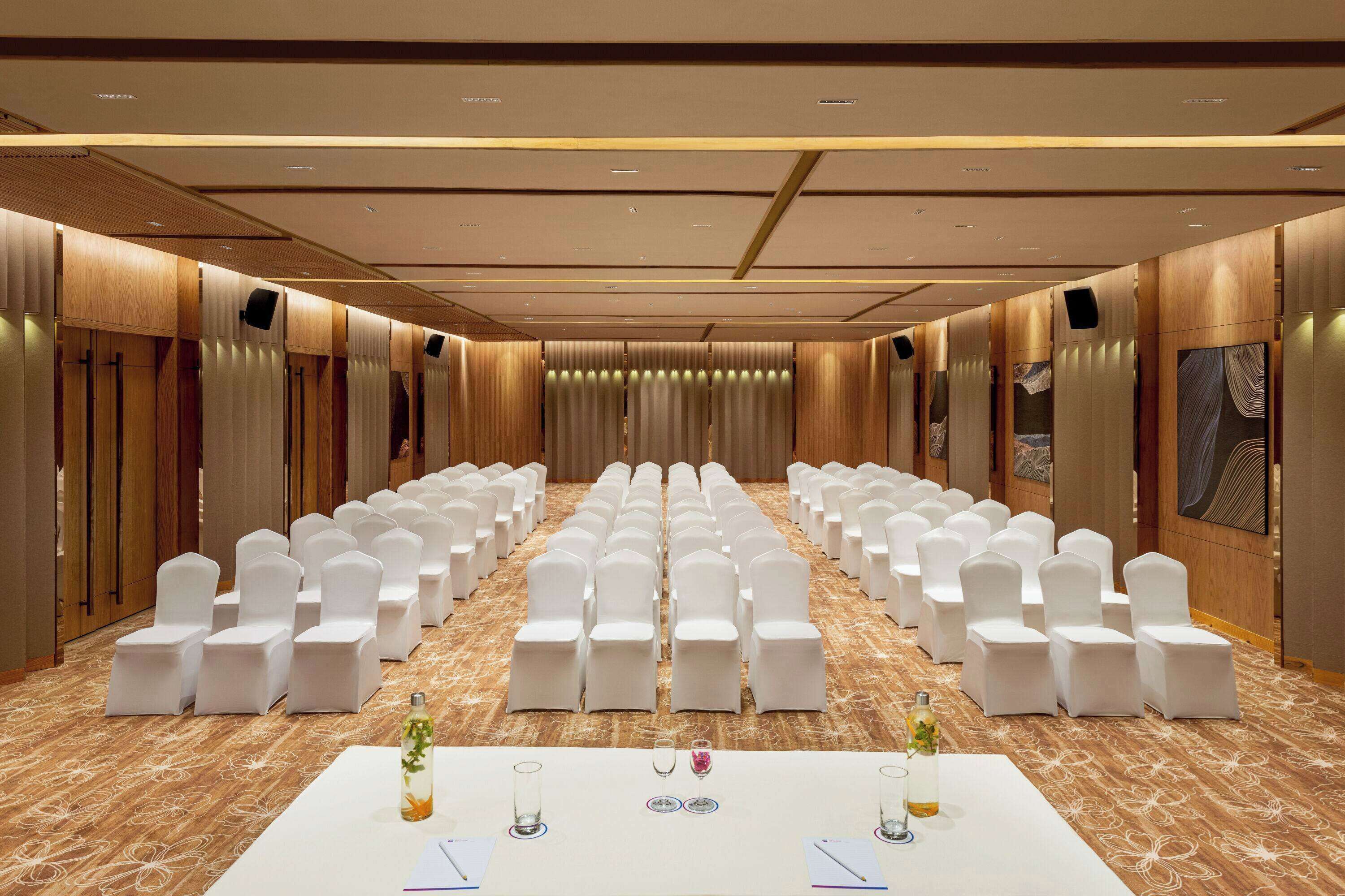 A conference room with rows of white chairs, a long table with nameplates, and a carpeted floor.