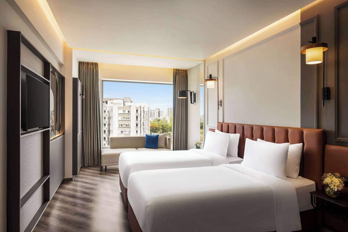 Quarto de hotel com duas camas, televisão, sofá, janela com cortinas e vista para a cidade.