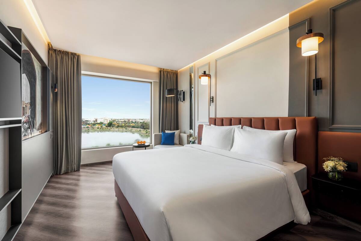 Quarto de hotel moderno com uma cama grande, vista para um lago e para a paisagem urbana.