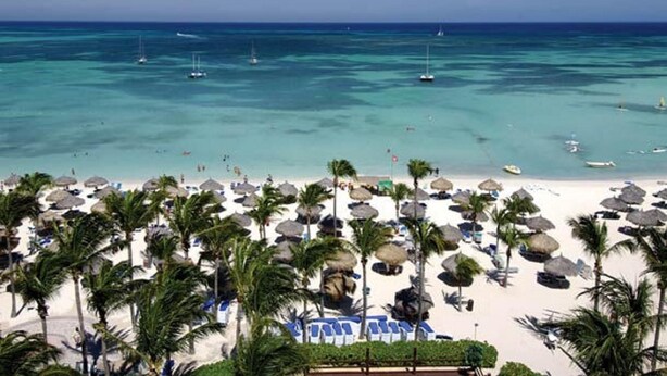 Una playa con aguas turquesas cristalinas, varios barcos y una hilera de palmeras y sombrillas.