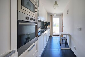 A narrow kitchen with a built-in oven, a gas stove, and a small dining table with a stool.
