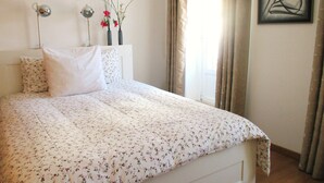 Une chambre à coucher avec une couvre-lit à motifs floraux, une tête de lit blanche, une table de chevet avec deux lampes, un vase rempli de fleurs et une œuvre d’art encadrée accrochée au mur.