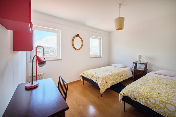 Habitación con dos camas, un escritorio con lámpara, radiador y ventana con vista a edificios.