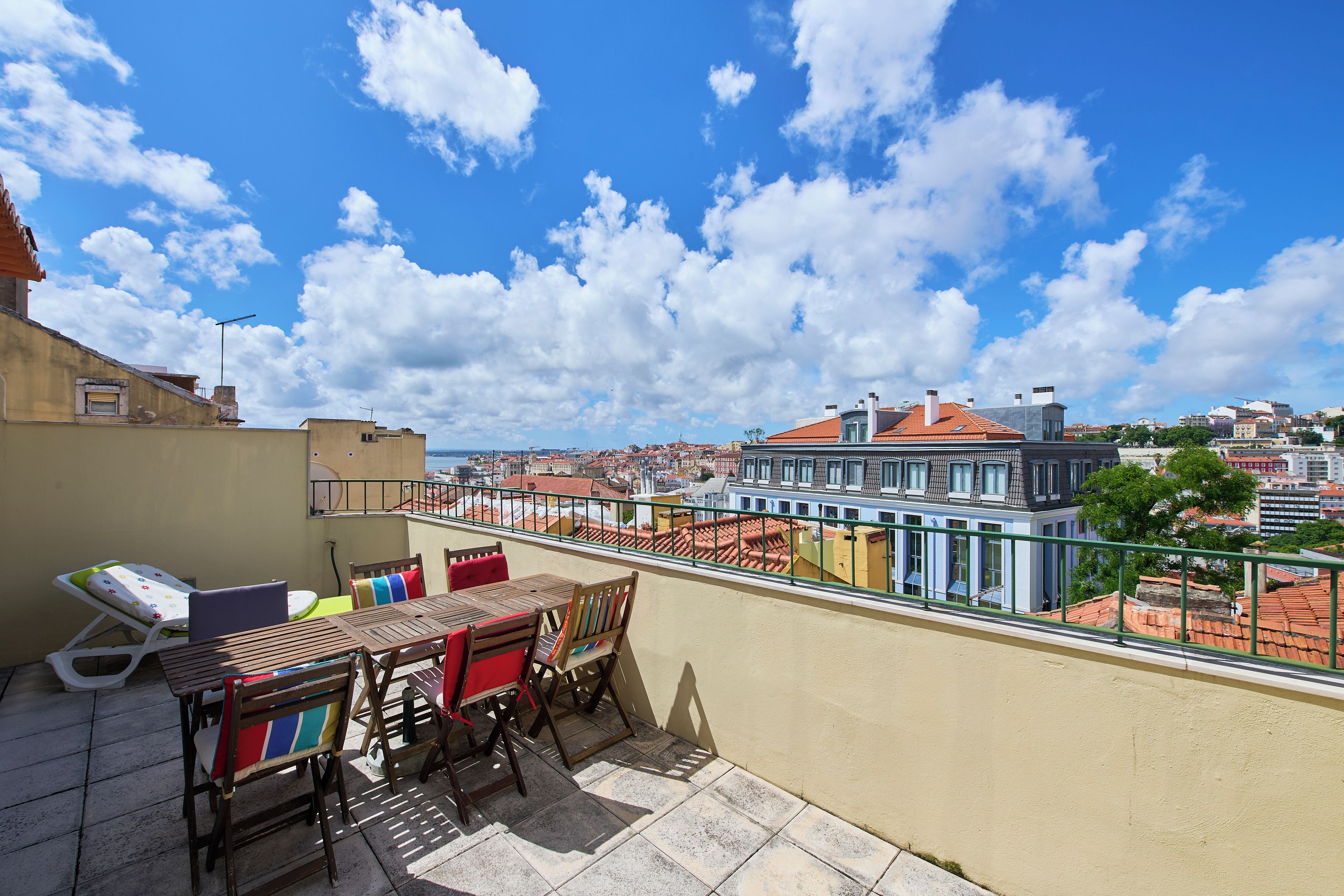 A rooftop terrace with a table and chairs, overlooking a cityscape with red-roofed buildings.