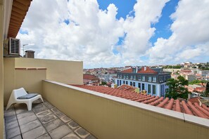 Un balcon donnant sur une vue panoramique de la ville, une chaise blanche et un toit recouvert de tuiles rouges.