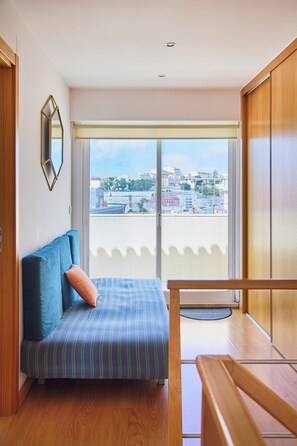 Una habitación con cama, un banco de madera, un ventanal con vistas a la ciudad y un espejo en la pared.