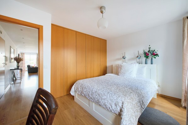 Ein Schlafzimmer mit Bett, Holzschrank, Nachttisch und Blick in den Wohnbereich.