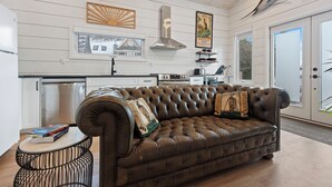 A modern kitchen with white cabinetry, a stainless steel range hood, and a dining area with a brown leather sofa and a small round table.