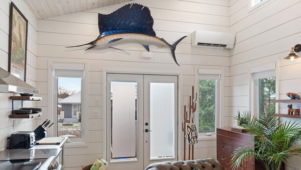 A modern kitchen with a large fish mounted on the ceiling, a dining area with a brown leather couch, and a wooden cabinet with a plant.