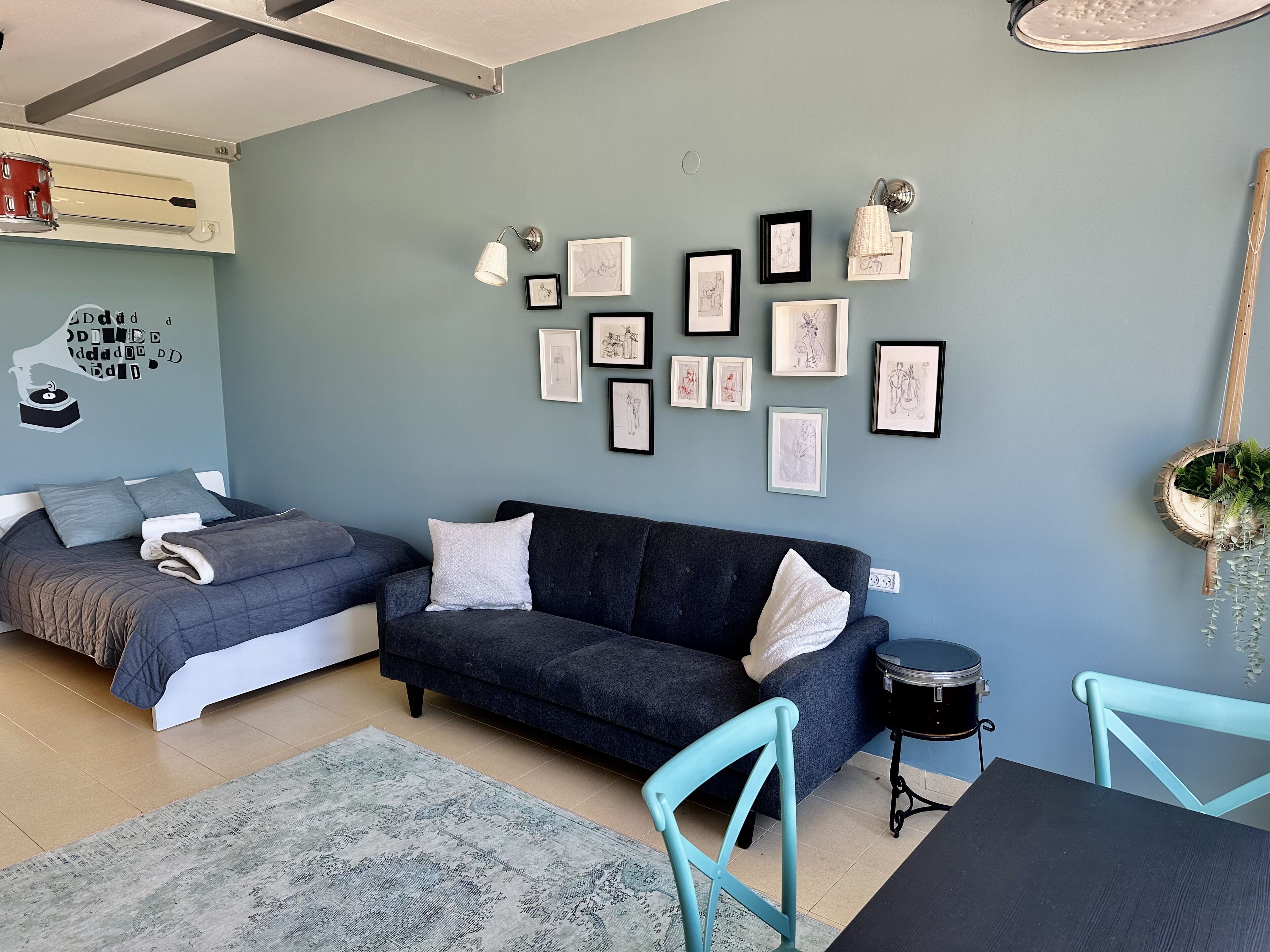 Ein modernes Wohnzimmer mit grauem Sofa, einem Bett mit grauer Bettwäsche und einem Esstisch mit zwei Stühlen. An der Wand befinden sich gerahmte Kunstwerke und auf dem Beistelltisch steht eine kleine Pflanze.