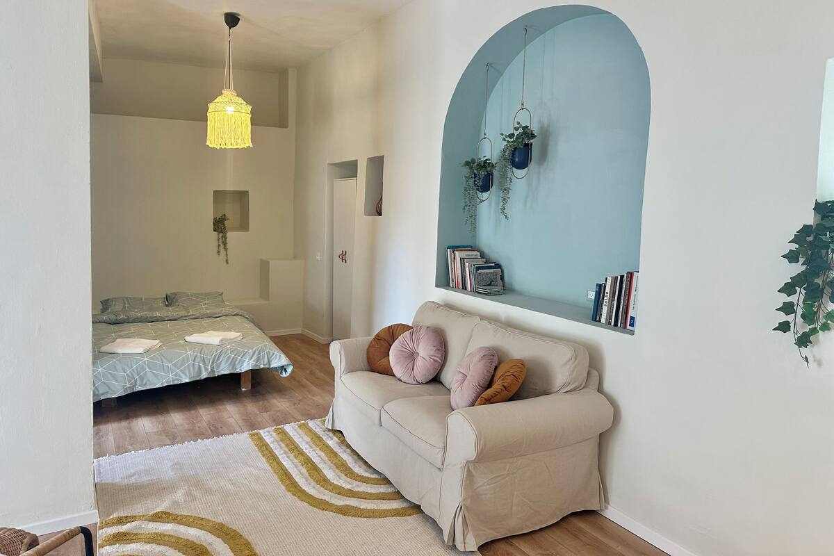 A modern living room with a sofa, a hanging light fixture, a bookshelf, and a large arched window.