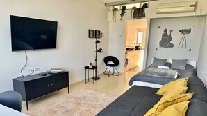 Ein modernes Schlafzimmer mit Bett, Schreibtisch, Fernseher und einer kleinen Küchenzeile im Hintergrund.
