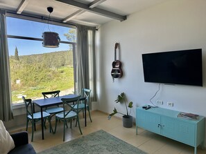 Un salon comprenant un coin repas, une guitare, un téléviseur et donnant sur un espace verdoyant.