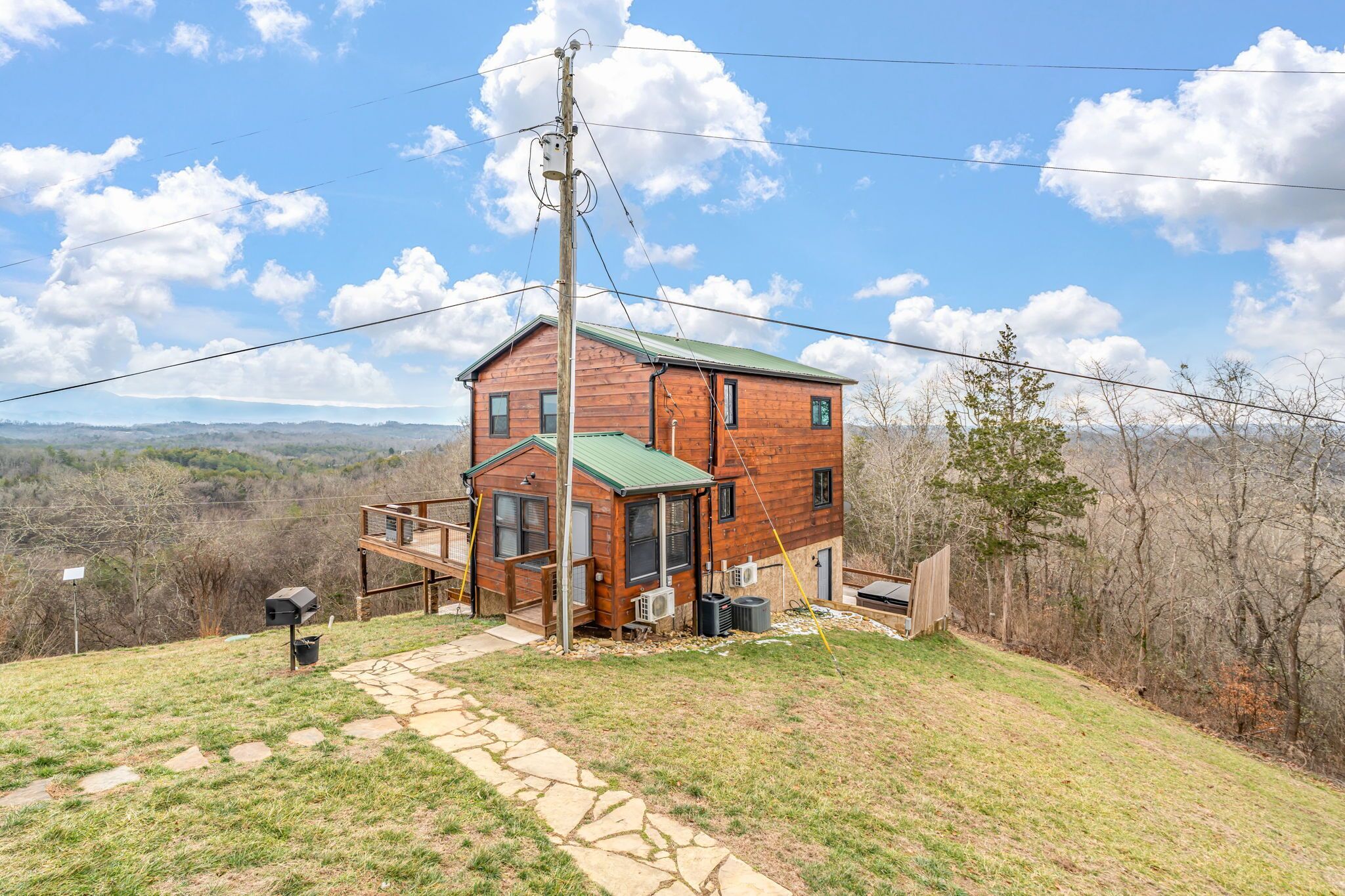 A two-story wooden cabin with a green roof, a stone pathway leading to the entrance, and a view of the surrounding landscape.