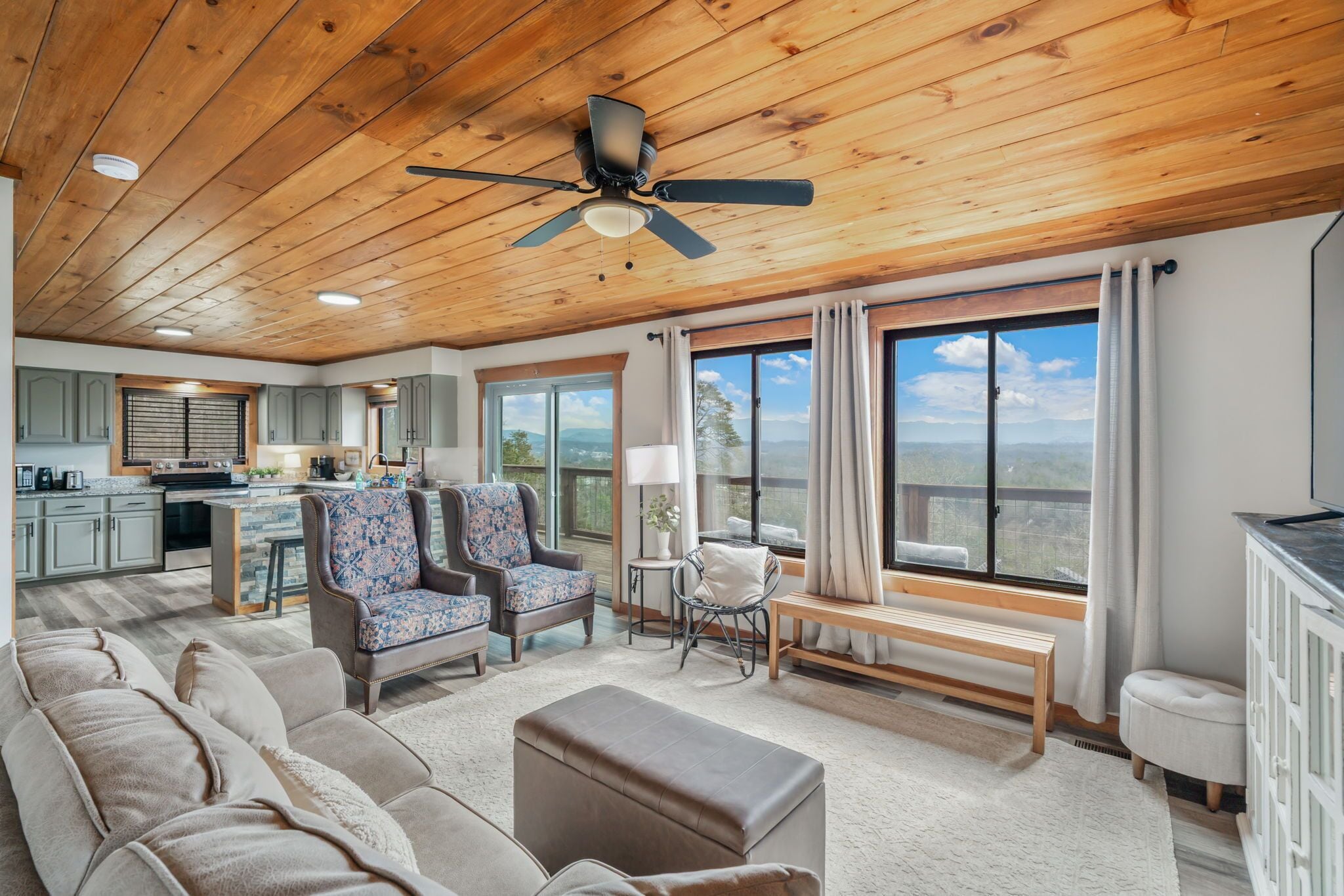 A spacious living area with a large window, a ceiling fan, and a kitchen in the background.