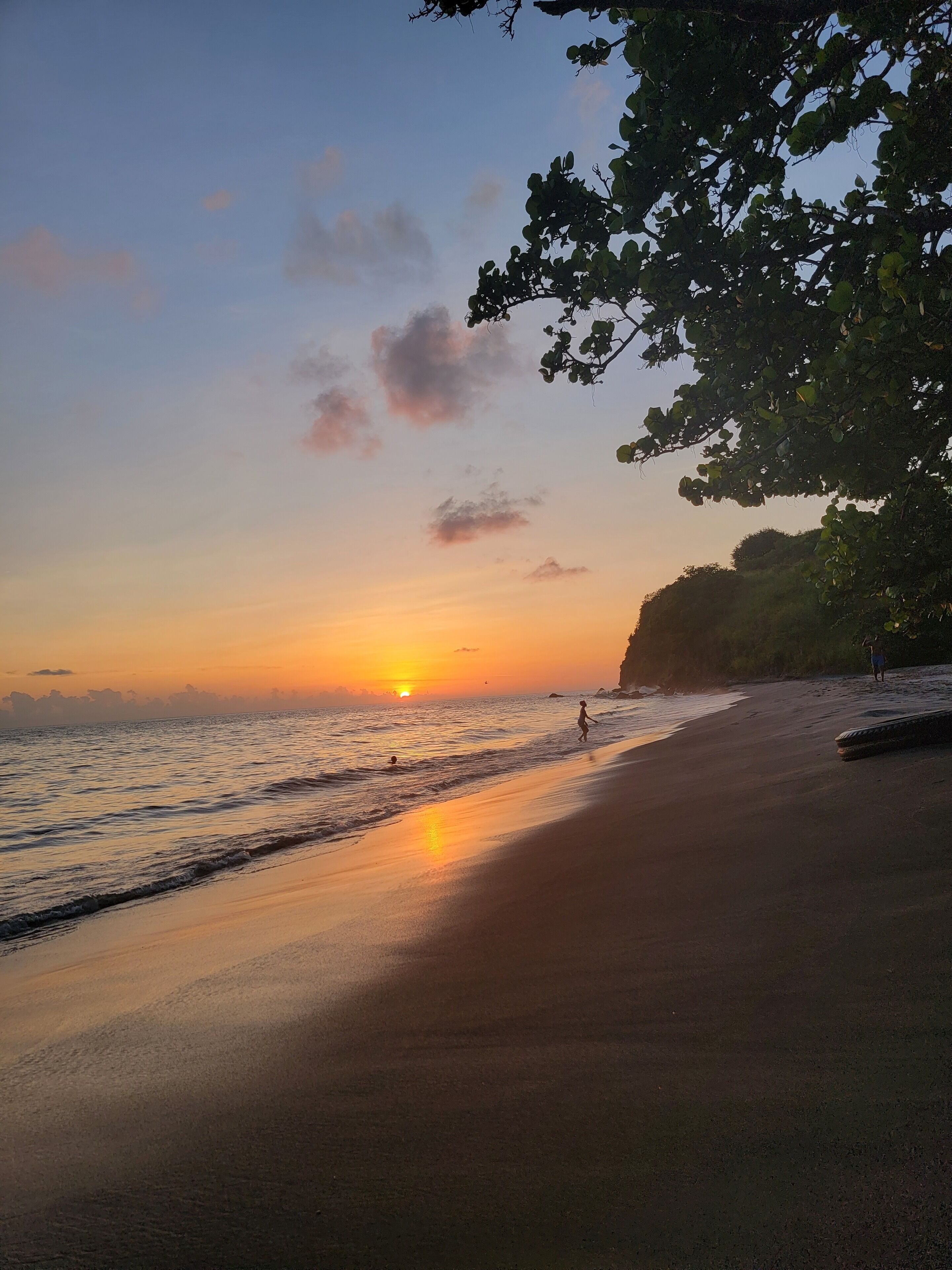 A beach at sunset with a clear sky, calm sea, and a few people walking along the shore.