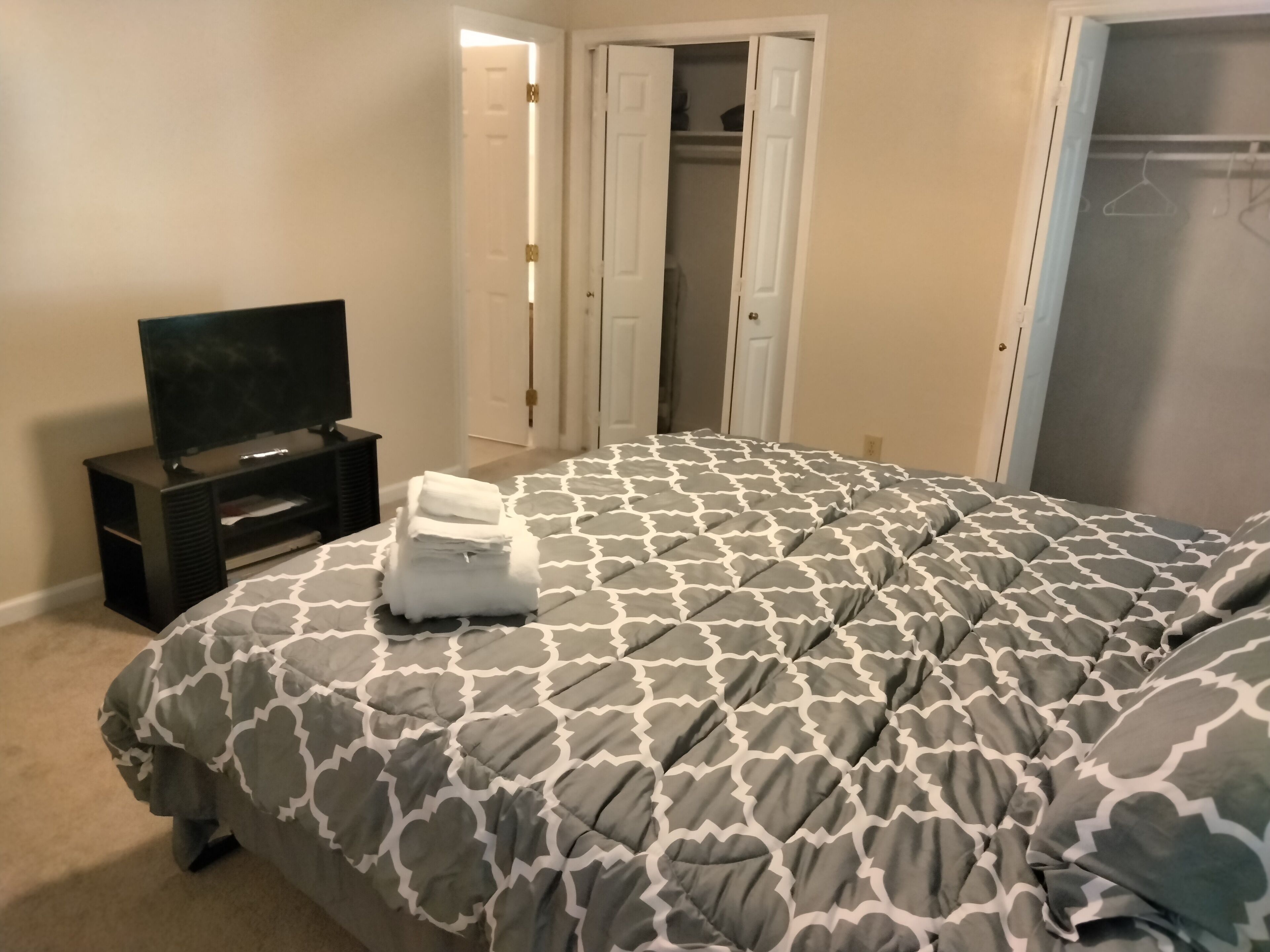 Um quarto com uma cama, uma televisão sobre um suporte e um guarda-roupa com portas abertas.