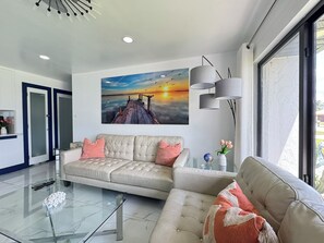 Un salon moderne avec un canapé beige, une table basse en verre et un tableau représentant un coucher de soleil sur l’eau.