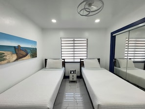 Une chambre d’hôtel avec deux lits simples, une petite table de chevet, une fenêtre avec des stores, un ventilateur de plafond et un tableau représentant une scène de plage accroché au mur.