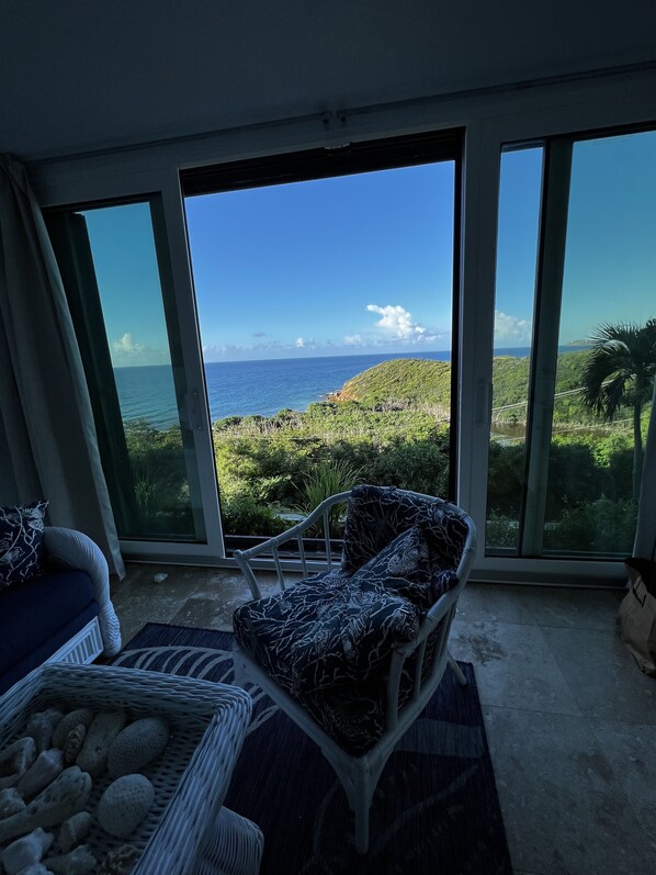 A room with a view of the ocean, a chair, a sofa, and a basket of items.