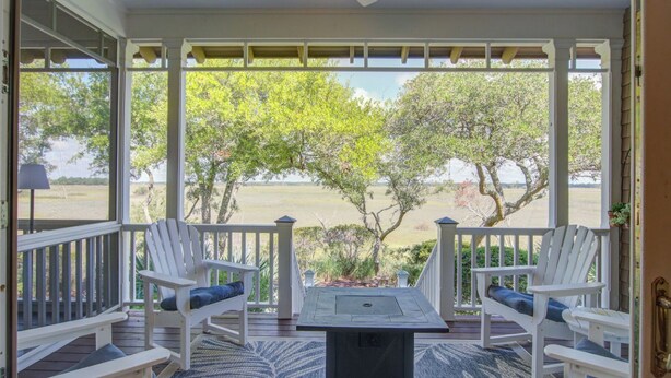 A covered porch with white railings, two Adirondack chairs with blue cushions, a table, and a view of a lake and trees. - Kiawah Exclusives | 65 Persimmon Court (Johns Island)