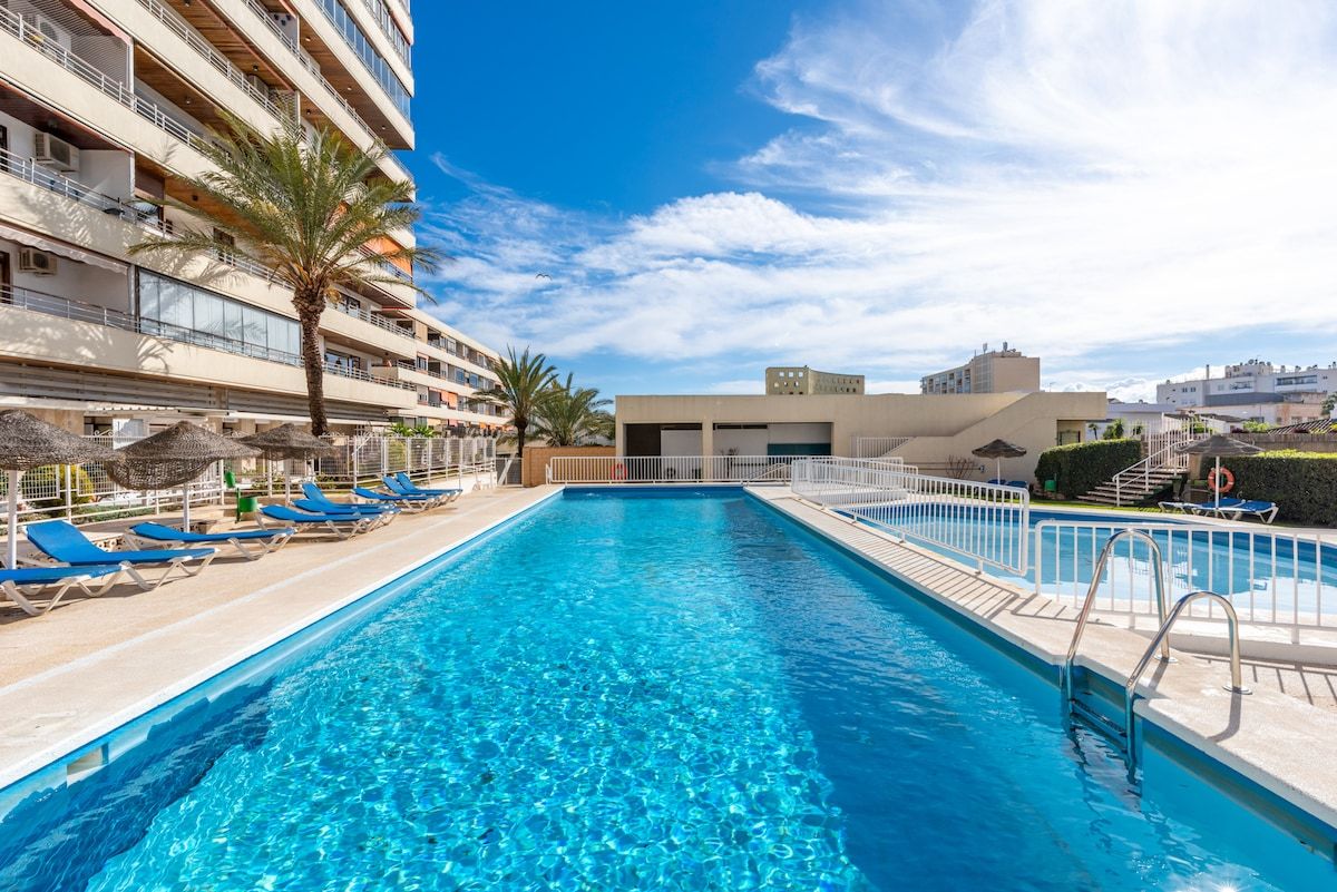 A swimming pool with lounge chairs and a palm tree, surrounded by a building with balconies.