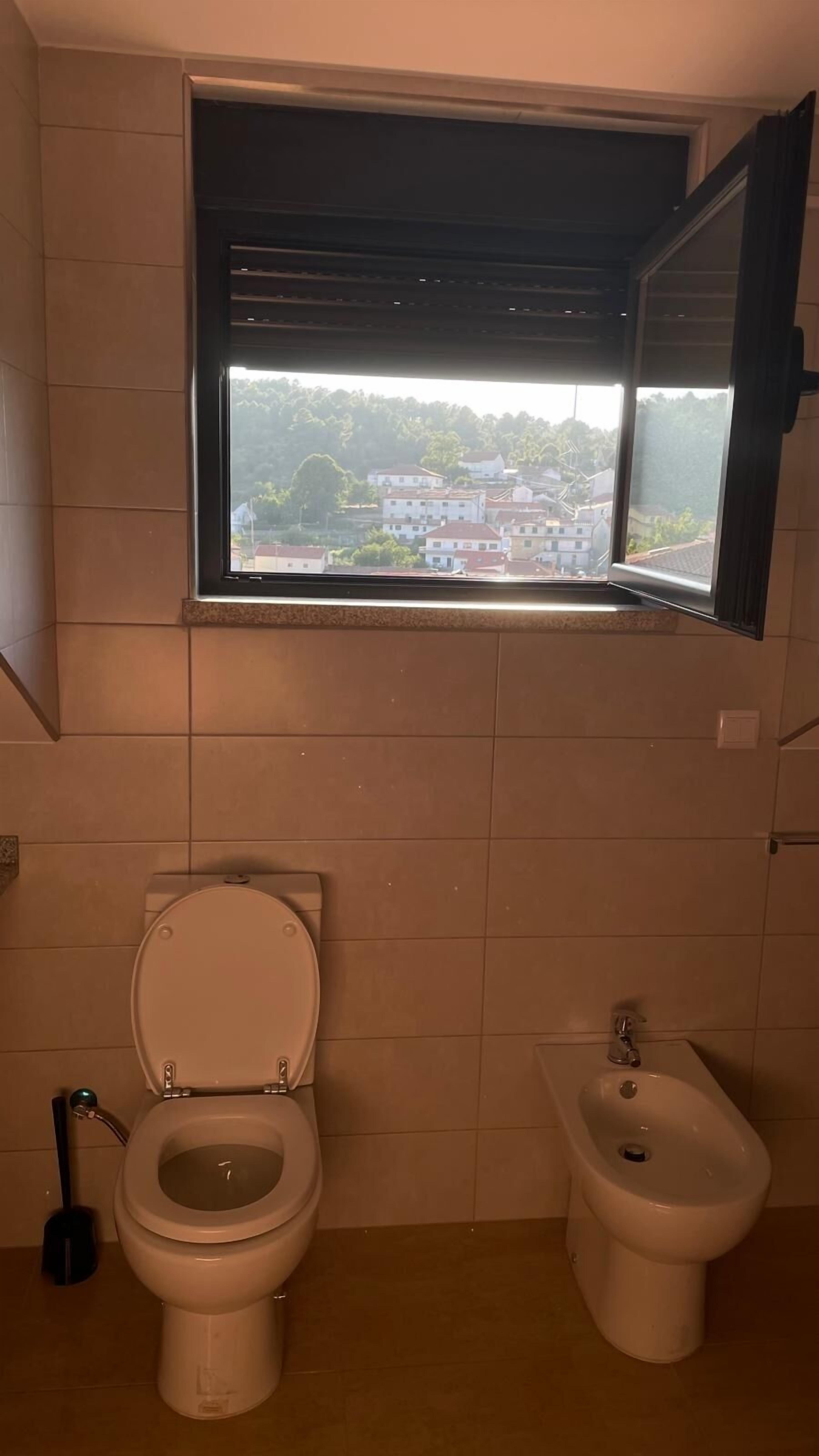 A bathroom with a toilet, bidet, and a window with a view of buildings and trees.