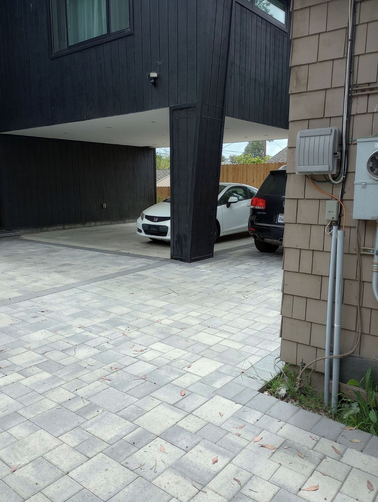 Een parkeerplaats met twee auto's, een modern gebouw met donkere gevelbekleding, en een stroomkast aan de rechterkant.