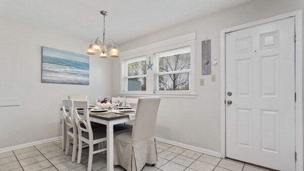 A dining area with a table set for four, white chairs, a chandelier, and a window with blinds. - Sandy Sands Retreat! Just 1 min to the beach! (Gulfport)